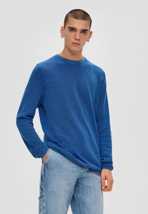 Blauwe gebreide trui met lange mouwen en ronde hals, gecombineerd met lichtblauwe jeans. Het model heeft kort haar en heeft de handen in de zakken.