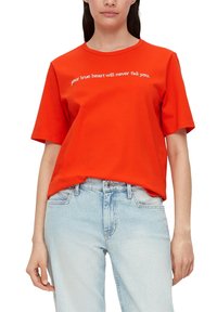 Rode katoenen t-shirt met korte mouwen en een ronde hals, voorzien van witte bedrukte tekst over de borst. Gepaard met lichtblauwe jeans.