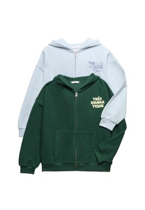 Twee opgevouwen hoodies met rits, één lichtblauw met de tekst "The World Is Your Oyster", één donkergroen met de tekst "Très Dramatique" op de borst.