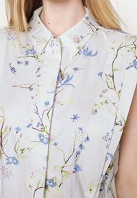 Light-colored sleeveless blouse featuring a floral pattern with blue and green accents. Collared design, smooth fabric with a soft texture.