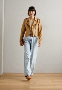 Tan leather biker jacket, beige top, high-waisted light blue jeans, brown belt, and brown sandals. Walking on wooden floor, gray background.