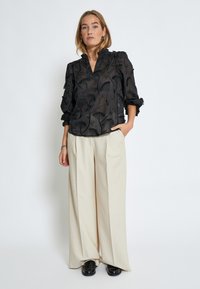 Blusa nera con ricami floreali, tessuto semi-trasparente, scollatura arricciata e maniche a sbuffo. Abbinata a pantaloni beige a gamba larga e scarpe nere.