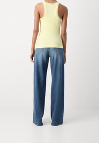 Person som bär en ärmlös topp med gul spetskant och löst sittande blå denimjeans, står vänd bort från kameran mot en enfärgad bakgrund.