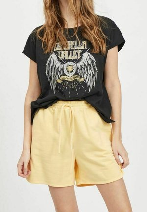 Sort grafisk t-shirt med vinge-design og tekst, kombineret med højtaljede gule shorts med elastik i taljen og snøre.