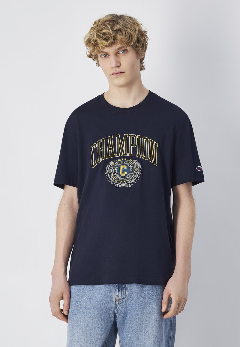 Camiseta de algodón de color navy con un gran logo y diseño de escudo "Champion" en múltiples colores. Mangas cortas, corte relajado, textura suave.