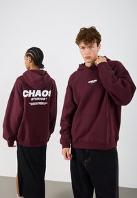 Hanorac maron cu logo alb "CHAOS STUDIOS". Dispune de o buzunar frontal tip pouch, glugă cu șnur și manșoane ribbate. Textură de țesătură netedă.