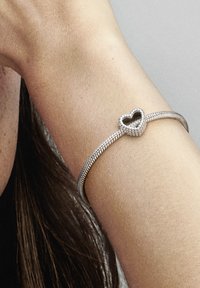 Pulsera de plata con un centro en forma de corazón, que presenta un diseño trenzado y una textura suave, llevada en una muñeca.