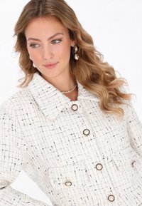 Biała marynarka z tweedu o teksturowanym wzorze, z kołnierzem, czterema kieszeniami i dużymi guzikami w perłowym wykończeniu.