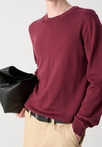 Burgunderfarbener Strickpullover mit langen Ärmeln und Rundhalsausschnitt. Kombiniert mit beige Hosen und einer schwarzen Lederhandtasche, die in einer Hand gehalten wird.