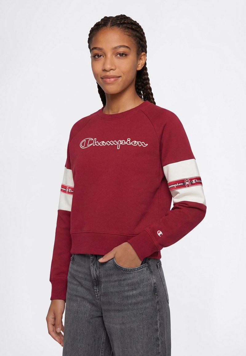 Jeune femme aux cheveux tressés portant un sweat-shirt rouge Champion avec des bandes blanches et rouges sur les manches et un jean gris, main dans la poche.