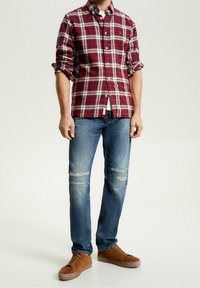 Homme portant une chemise à carreaux rouges à manches retroussées, un jean bleu délavé et des baskets en daim marron, se tenant contre un fond uni.