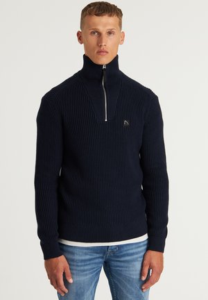 Marineblaue Rippstrickpullover mit hohem Kragen und teilweise Reißverschluss vorne. Mit einem kleinen Logo-Detail auf der Brust und engen Bündchen.