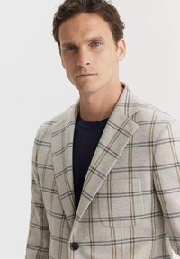 Jasnoszary marynarka w kratę, wykonana z teksturowanej tkaniny. Posiada klapy w kształcie notch, pojedynczy guzik oraz kieszeń naszywaną z brązowymi akcentami.