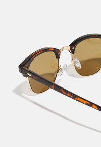 Schildpatt-Sonnenbrille mit goldenen Akzenten, ovalen Gläsern und klaren Nasenpolstern. Die Bügel zeigen ein dunkles Muster und ein dezentes Markenlogo.