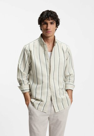 Homme portant une chemise beige clair à rayures verticales avec les manches retroussées et un pantalon de couleur claire, mains dans les poches, debout devant un fond blanc.