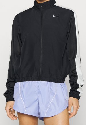 Femme portant une veste noire zippée avec des rayures blanches sur les manches et un short de sport violet clair.