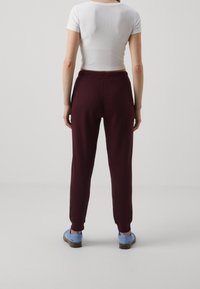 Burgunderfarbene Jogginghosen mit lockerer Passform, elastischem Bund und Knöchelbündchen, kombiniert mit einem weißen T-Shirt mit kurzen Ärmeln und blauen Schuhen.