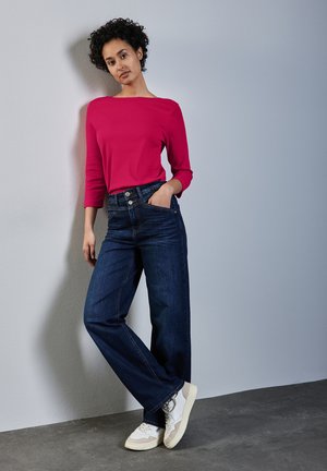 Rotes, geripptes Langarmtop kombiniert mit dunkelblauen, weiten Jeans. Weiße und beige Sneaker mit strukturiertem Design runden das Outfit ab.