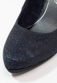 Gros plan d'une chaussure plate bleu foncé pailletée avec un bout arrondi et une surface texturée sur fond blanc.