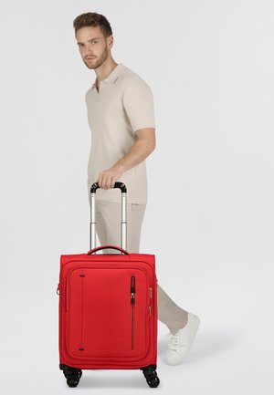 Maleta roja con una tela texturada, bolsillo con cremallera, asa retráctil y cuatro ruedas, posada al lado de una persona vestida con ropa clara.