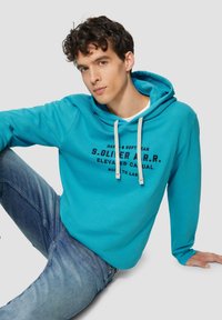 Türkise Kapuzenjacke aus weichem Material mit Kordelzug und bedrucktem Logo. Kombiniert mit hellblauen Jeans, Modell sitzt vor einem neutralen Hintergrund.