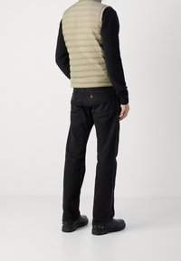 Smanicato imbottito beige con cuciture orizzontali, indossato sopra una camicia nera a maniche lunghe, abbinato a jeans neri e scarpe nere.