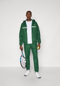Green tracksuit made of smooth fabric, featuring white accents and logos. The person holds a tennis racquet and ball, wearing white sneakers.