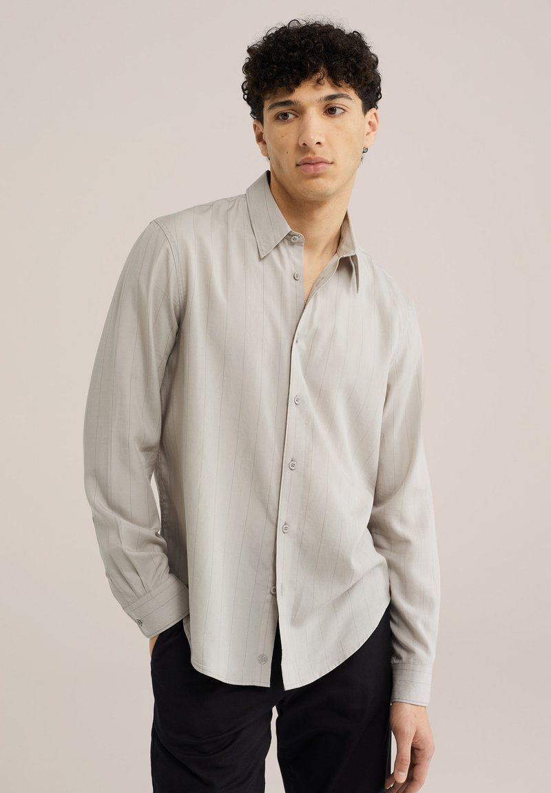 Jeune homme aux cheveux bouclés portant une chemise beige clair à manches longues boutonnée et un pantalon noir, debout devant un fond uni.