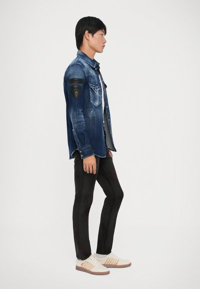 Giacca di jeans di colore blu scuro con accenti sbiaditi e toppe in pelle nera, abbinata a pantaloni neri aderenti e sneakers bianche con motivi a righe.