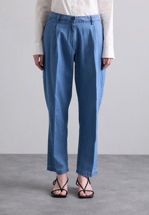 Person trägt hellblaue, weit geschnittene Hose und schwarze Sandaletten mit Riemchen, steht auf einem grauen Boden vor einem schlichten grauen Hintergrund.