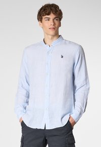 Giovane uomo che indossa una camicia leggera azzurro chiaro con colletto alla coreana, in piedi con sicurezza con le mani in tasca su uno sfondo chiaro.