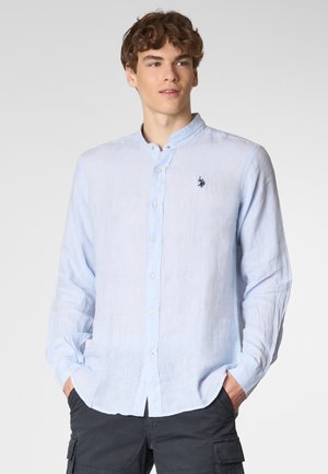 Giovane uomo che indossa una camicia leggera azzurro chiaro con colletto alla coreana, in piedi con sicurezza con le mani in tasca su uno sfondo chiaro.