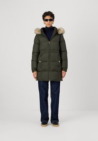 Tumši zaļš pūkains jaka ar mākslīgā kažoka kapuci, ar rāvējslēdzēju un divām priekšējām kabatām. Uzvilkts virs tumši zilas kreklas un tumšiem džinsiem.