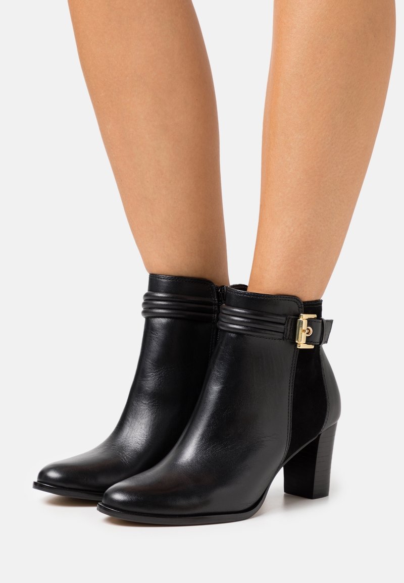 Bottines en cuir noir avec talons carrés, sangles décoratives et boucle dorée, portées sur des jambes nues.