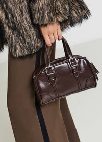 Sac à main en cuir marron avec de courtes poignées, en forme rectangulaire, avec des coutures visibles et des détails de boucle. Porté avec un manteau à texture en fourrure et un pantalon marron.