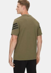 T-shirt verde oliva a maniche corte con tre strisce nere sulla spalla sinistra, collo rotondo e design aderente. Tessuto in cotone liscio.