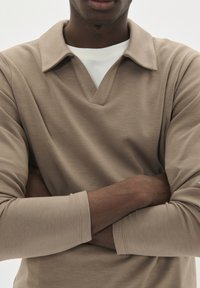 Hombre con los brazos cruzados, vistiendo una camisa de manga larga beige sobre una camiseta blanca de cuello redondo, frente a un fondo claro y liso.