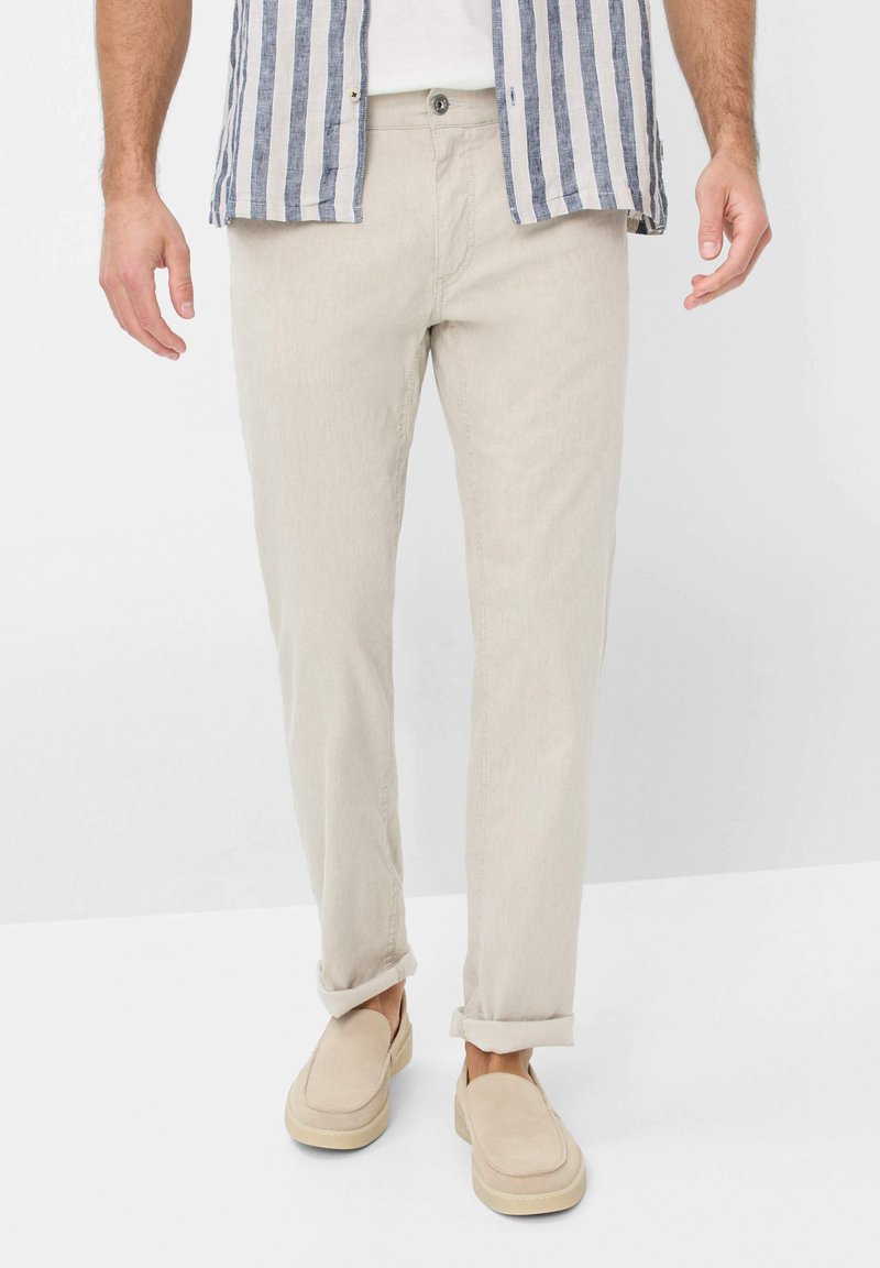 Beige lightweight linen pants with a straight fit, paired with a white T-shirt and striped blue and white button-up shirt, worn with slip-on shoes.
