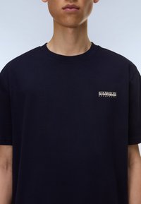 Noor inimene kannab musta Napapijri Geographic T-särki, millel on vasakul rinnal logo, taust on ühtlaselt hele.