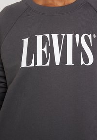 Šedá mikina s vláknitou strukturou a bílým logem "LEVI'S". Má klasický kulatý výstřih a raglánové rukávy pro uvolněný střih.