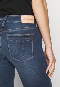 Jeans de mezclilla azul oscuro con un parche de cuero que dice "CALVIN KLEIN JEANS" en la cintura y costuras distintivas en los bolsillos traseros.