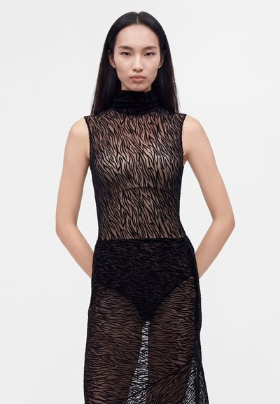 Femme aux longs cheveux noirs portant une robe noire transparente sans manches à col haut avec des motifs semblables à ceux du zèbre et des sous-vêtements noirs.