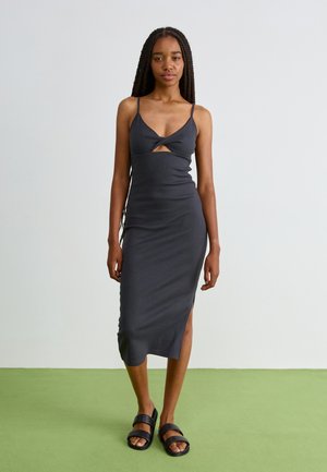 Robe midi noire sans manches avec décolleté en V et découpe à l'avant, présentant une coupe ajustée et une fente sur le côté, associée à des sandales à glissière noires.