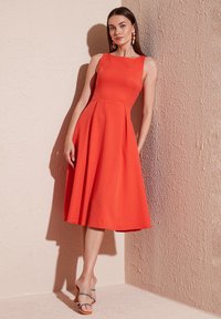 Röd ärmlös klänning med en figursydd bodice, utsvängd kjol och slät textur. Kompletterad med remsandaler i flera färger.