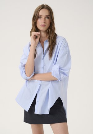 Light blue button-up shirt with rolled sleeves, made of smooth fabric. Features a curved hem and relaxed fit. Worn over a black pinstriped skirt.