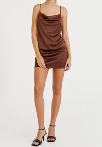 Robe nuisette en satin marron avec fines bretelles, design drapé et ourlet court. Associée à des sandales à talons noires avec brides autour de la cheville.