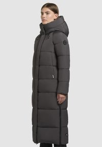 Lange, puffy grijze winterjas met een hoge kraag en verwijderbare capuchon. Voorzien van horizontale stiksels, een voorrits en zijzakken.
