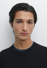 Junger männlicher Model mit dunklem Haar, trägt einen strukturierten schwarzen Pullover über einem blauen Hemd. Neutraler Hintergrund, Fokus auf die Gesichtszüge.