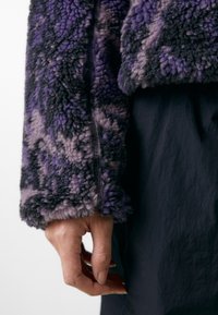 Giacca in pile con un motivo astratto in viola, nero e grigio; presenta un'apertura con zip e un tessuto testurizzato.