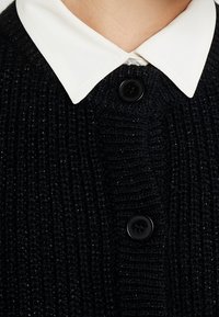 Schwarzer gestrickter Pullover mit strukturiertem Muster, ausgestattet mit einem weißen, flachen Kragen und drei schwarzen Knöpfen auf der Vorderseite.